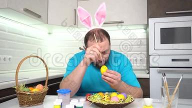 复活<strong>节</strong>快乐。 一个穿着兔子耳朵蓝色t<strong>恤</strong>的男人在厨房里画准备复活<strong>节</strong>的鸡蛋。