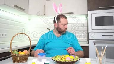 复活节快乐。 一个穿着兔子耳朵蓝色t恤的男人在厨房里画准备复活节的鸡蛋。