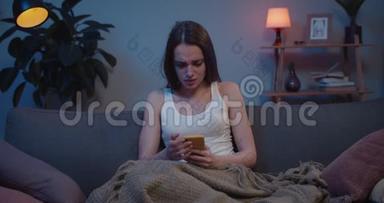 女孩坐在沙发上用智能手机和思考突发问题的正面视图。 年轻悲伤的女人捂脸