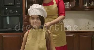 戴着厨师帽的白人女孩微笑着在厨房跳舞，她的母亲在她身上系着围裙。 幸福的特写肖像