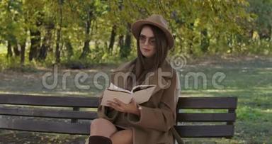 聪明优雅的高加索女人戴着太阳镜和棕色帽子坐在长凳上翻书页。 美丽美丽