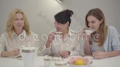 三个不同年龄的白人妇女坐在桌子旁喝茶或咖啡。 聚集的朋友或家人