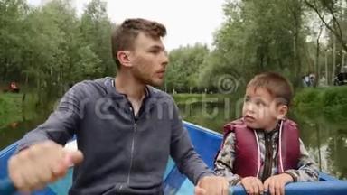 年轻快乐的父亲驾驶木船，儿子帮助他。父亲和儿子在船上划船，在我的绿色公园里互相交谈
