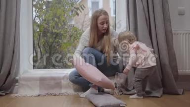 年轻迷人的白人女人坐在低矮的窗台上和小女孩说话。 穿着粉红色衬衫走路的可爱孩子