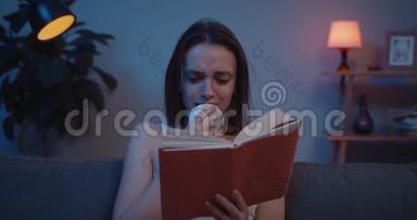 年轻沮丧的女人坐在沙发上，读着非常悲伤的书。 情感女孩在书中的情节和情节中担心戏剧时刻