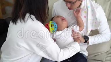 年轻的亚洲父亲坐在地板上，<strong>抱着婴儿</strong>，而母亲则拿<strong>着婴儿</strong>衣服，一起照顾孩子