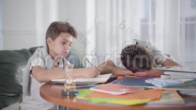 聪明的白人男孩做家庭作业的肖像，他的兄弟睡在桌子上。 孩子醒来时他的兄弟姐妹