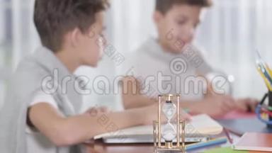 当两个模糊的小学生在背景下画画或写作时，旋转沙漏的特写停留在桌子上。 双胞胎