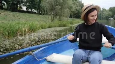 年轻的快乐女孩戴着复古的帽子，漂浮在蓝色的木船上，湖面上挂着睡莲。 害羞的女孩在城市公园的湖面上划桨。 S.S.