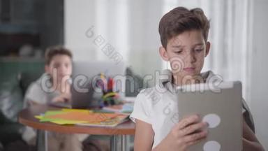 用平板电脑作为他的孪生兄弟坐在笔记本电脑的背景下的白种人男孩的特写肖像。 小兄弟