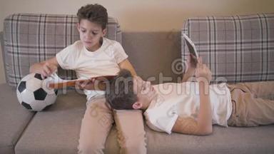 白人男孩坐在沙发上，拿着笔记本和球，他的孪生<strong>兄弟</strong>跪着，用平板电脑。 <strong>兄弟</strong>姐妹
