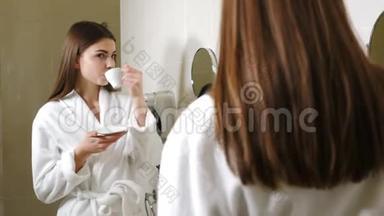 年轻迷人的女人穿着<strong>白色浴袍</strong>在<strong>浴</strong>室镜子前喝咖啡。 后视镜头。 美丽的女人