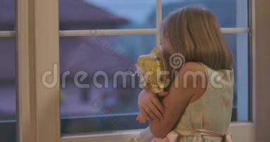 吓坏了的白人金发女孩拥抱娃娃，看着窗外。 孤独的孩子害怕黑暗拿着玩具