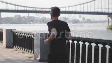 活泼苗条的男人穿着黑色t恤和运动短裤沿着河跑。 年轻的职业运动员在城市跑步。 健康强