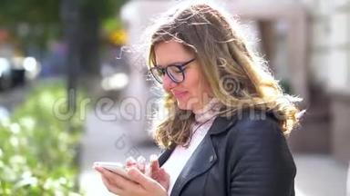 一个戴着眼镜的漂亮女孩站在树上，带着智能手机，微笑着和它一起工作。 美丽的人。