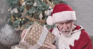 圣诞老人的特写镜头坐在圣诞树的背景下，摇着礼盒。 好奇的老头