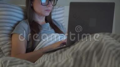 戴眼镜的女人晚上躺在床上用笔记本电脑工作。 移动成瘾或<strong>失眠</strong>的概念。