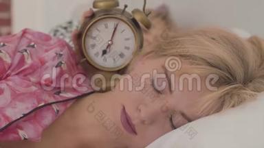 一位美丽的金发白种人女人正在睡觉，她的朋友正在接近闹钟，闹钟显示她点