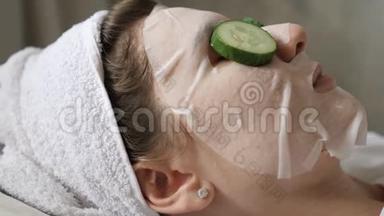 用新鲜黄瓜和保湿面膜进行面部美容治疗。 头上戴着白色毛巾的女人