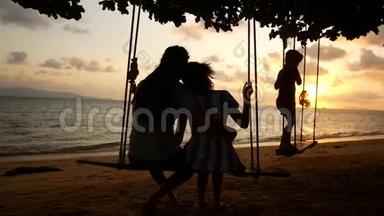 度假的幸福家庭。 爸爸妈妈和宝宝骑在秋千上，看海边的日落