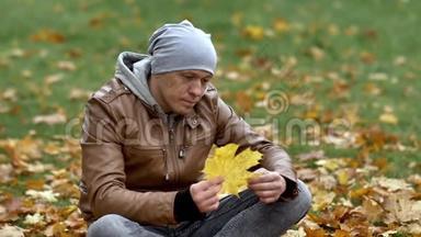 一个年轻人坐在秋天公园的草地和黄色的枫叶上。 仔细检查一大片枫叶