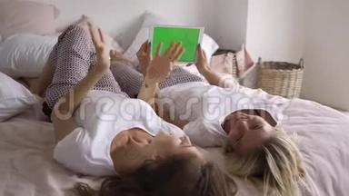 两个穿着白色t<strong>恤衫</strong>的年轻漂亮女朋友躺在床上，用绿色屏幕观看平板电脑上的信息