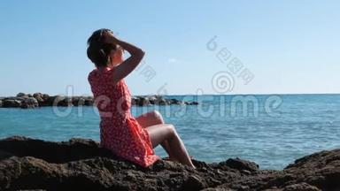 年轻苗条的白种人女孩穿着红色短裙坐在大海上的大石头上。 有魅力的黑发女人<strong>直发</strong>坐在r附近