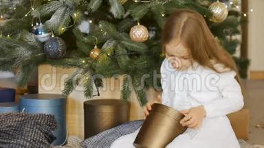 <strong>可爱</strong>的金发女孩打开了一个暗金<strong>礼盒</strong>，她坐在圣诞树旁，快乐的童年和圣诞节