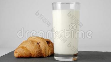 新鲜的白牛奶放入透明的玻璃中，靠近牛角面包，巧克力在白色桌子上的黑板上旋转
