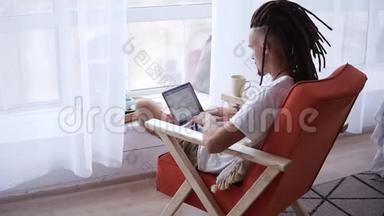 一个穿着白色T恤、戴着深色骷髅的男人坐在客厅里用笔记本<strong>电脑</strong>工作的<strong>背面</strong>镜头