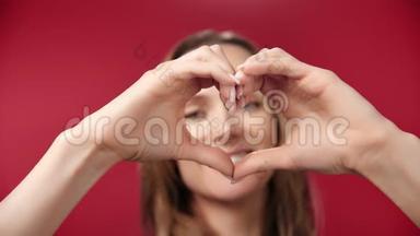 近距离微笑的年轻女子在红色摄影棚背景下用手摆姿势做心形标志