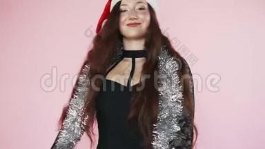 美丽的红发女孩戴着圣诞老人的帽子，旋转着黑色的连衣裙。 新年的心情。 圣诞节。 慢动作粉红色背景关闭。