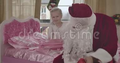 惊讶的白种人女孩坐在粉<strong>红色</strong>的床上，微笑着作为圣诞老人穿着<strong>红色</strong>的服装，看着礼品袋。 <strong>儿童</strong>