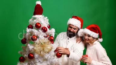 一对戴着红色圣诞帽的夫妇在绿色背景上装饰圣诞树。 家庭、圣诞节、寒假和