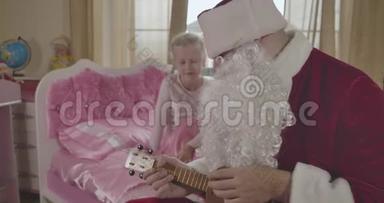 可爱的高加索儿童坐在粉红色的床上，看着圣诞老人玩四弦琴。 穿圣诞老人服装的成年男子