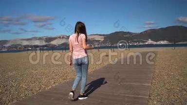 快乐迷人的女孩穿着粉红色的t恤和蓝色牛仔裤，或者女人在海滩上走在木路上，用智能手机聊天。