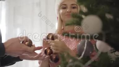 一幅美丽的白种人女人帮助无法辨认的男人在圣诞树上挂装饰品的肖像。 幸福的妻子