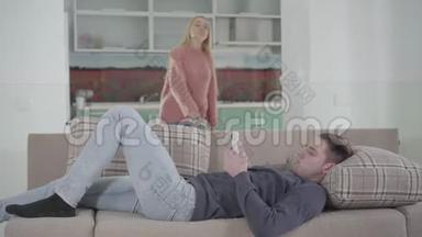 年轻的高加索男人躺在沙发上用智能<strong>手机</strong>的肖像，而他不幸的妻子在<strong>后台</strong>抽真空