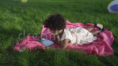 一个非洲裔小女孩躺在公园里，躺在床上，在平板电脑上看动画片