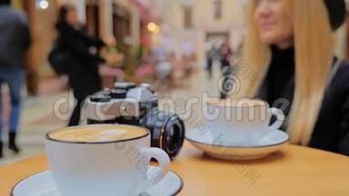 一个<strong>未被</strong>认出的白人女人，一个金发女郎坐在咖啡馆的桌子旁。 有两杯卡布奇诺咖啡和一杯