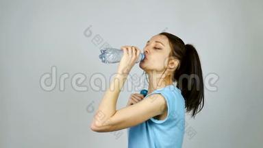 一个长着深色头发的布鲁内特女孩在灰色的环境下锻炼后，正在用塑料透明瓶子喝纯净水