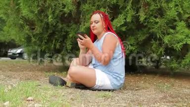 一个扎着红辫子的年轻女人正坐在草地上的公园里看着她的手机。 移动摄像机