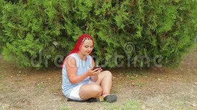 一个扎着红辫子的年轻女子坐在草地上的公园里看着她的手机