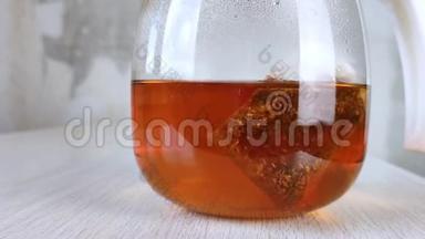 袋红茶用玻璃茶壶冲泡.. 木桌上透明圆罐中的棕色琥珀饮料特写。 混凝土墙