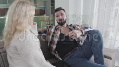 一个英俊的白种人男人的特写，黑发和胡须坐在沙发上和金发女人说话。 穿休闲服的男人