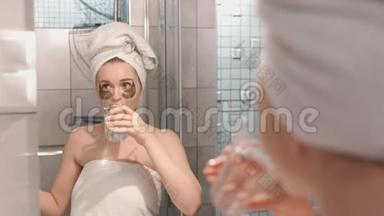 一幅美丽的年轻女子的肖像画，她穿着浴袍和毛巾，站在浴室里，眼睛下面有补水补丁，还有滴滴。