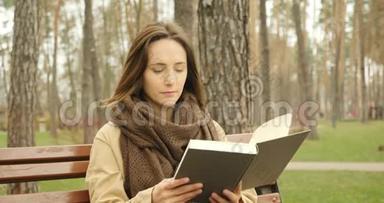可爱专注的女人翻书页，独自坐在寒冷的秋天公园的长凳上，戴着棕色的围巾看书。