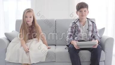 两个高加索儿童坐在沙发上的肖像。 在平板电脑屏幕上看东西的男孩，穿着<strong>漂亮衣服</strong>的女孩