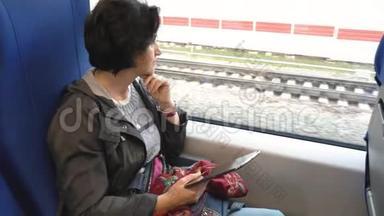 白种人女人坐在火车窗口，拿着平板电脑，看着窗外，支撑着她的下巴。