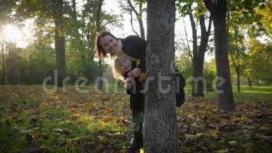 和妈妈一起玩，一个迷人的微笑女孩和她的父母一起在秋天的公园里玩，一家人躲在树干后面